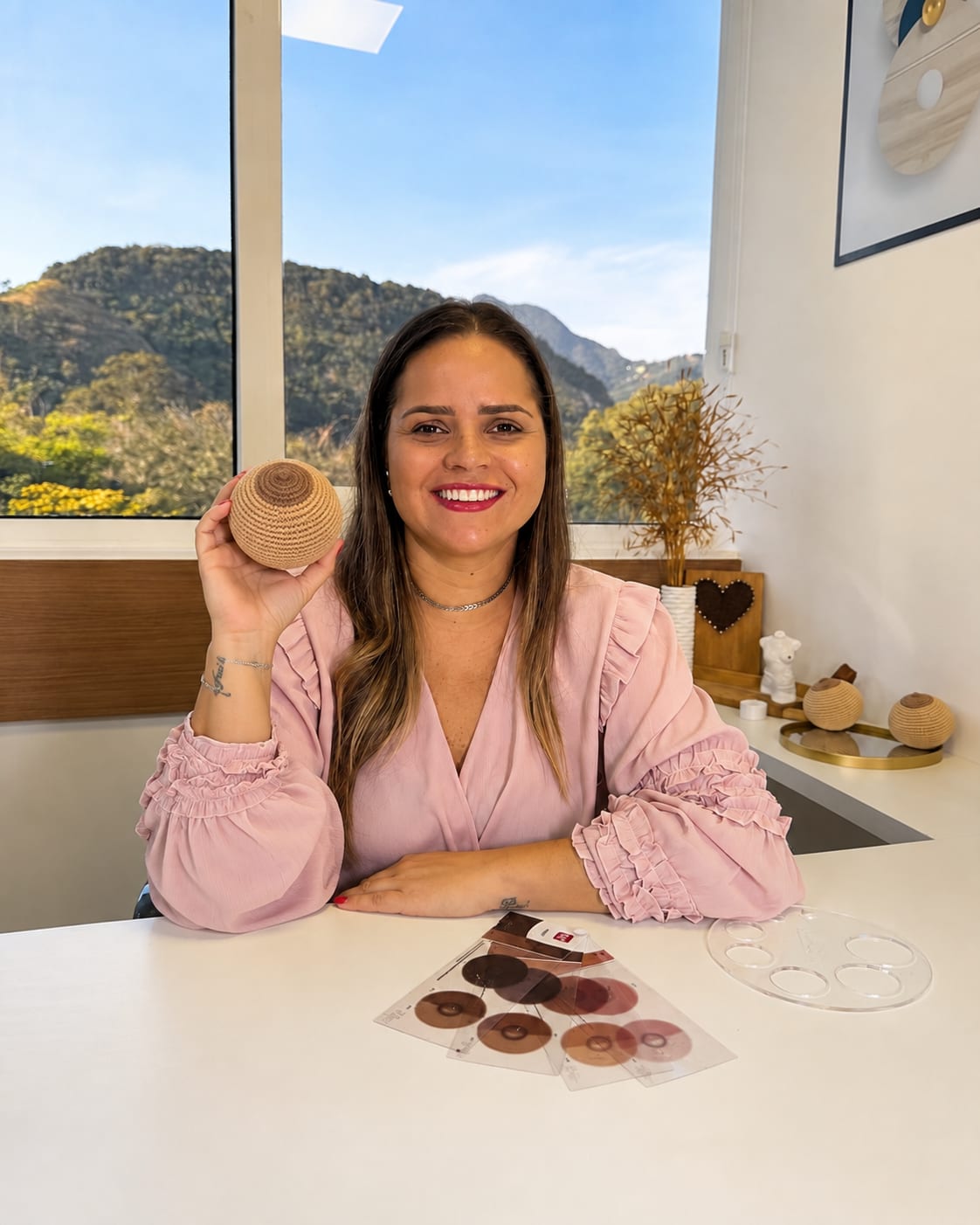 Carolina Vinuso sorrindo em seu consultório, segurando um modelo de mama em crochê, com cartelas de pigmentos sobre a mesa